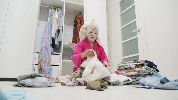 Wanita dengan masker wajah lembab melipat pakaian berserakan di lantai di kamar tidur — Stok Video