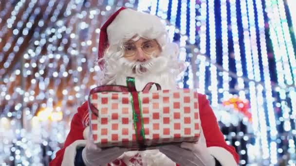 Jultomten ger en presentask på bakgrunden av ljusa julkransar — Stockvideo