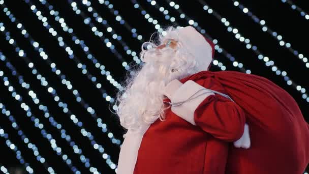 Santa Claus mira hacia arriba en el árbol de Navidad de la ciudad y luces intermitentes brillantes de guirnaldas — Vídeos de Stock