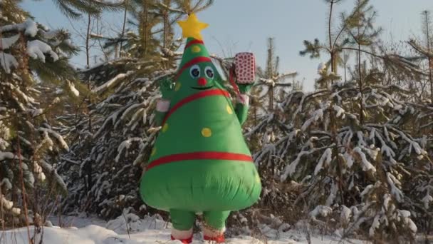 Un hombre en un disfraz de muñeco de árbol de Navidad está bailando alegremente con un regalo en su mano — Vídeo de stock