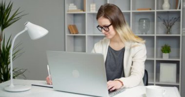 Genç kız öğrenci evde bilgisayar kullanarak ders çalışıyor ve bir not defterine yazıyor. Kız ev ödevini internette yapıyor. Uzak öğrenme