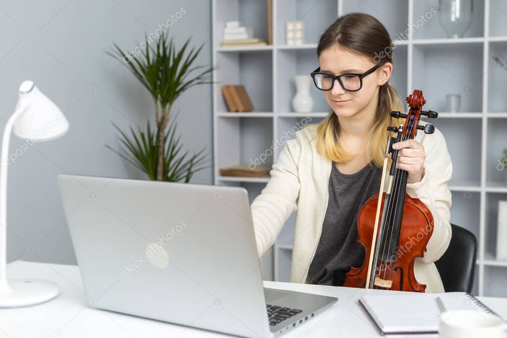 A girl plays the violin remotely online looking into a laptop. Music lesson at home by video call.