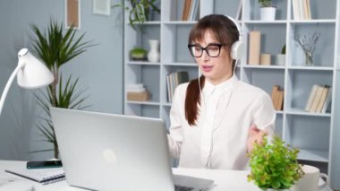 Kulaklıklı, dost canlısı genç bir bayan dizüstü bilgisayarla konuşuyor ve ona bakıyor. Webinar 'ın video görüşmesinde konuşan mutlu kız. Bir kız öğrenci internette ders çalışıyor..