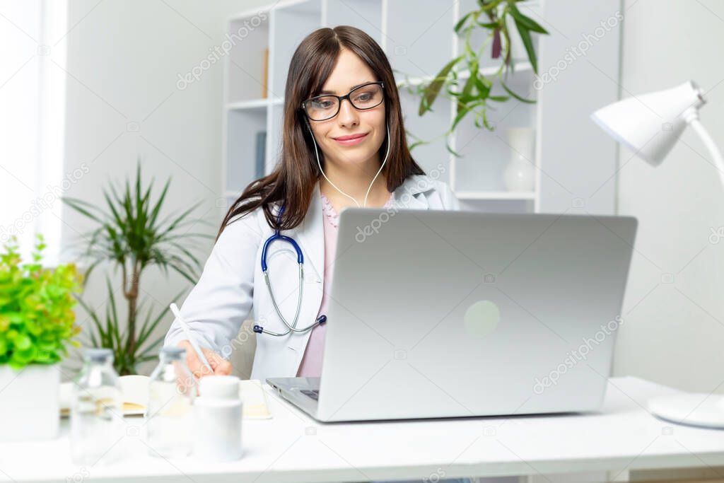 female therapist working remote, sitting at laptop with earphones and writing in notebook