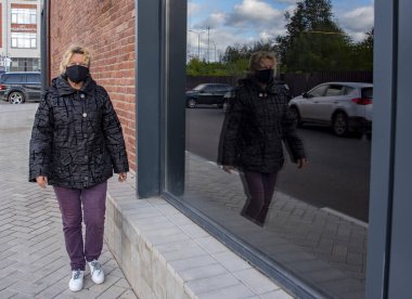 Rusya, Ruza, Eylül 2020. Siyah koruyucu maskeli yaşlı bir kadın caddede yürüyor ve bir binanın camından yansıyor..