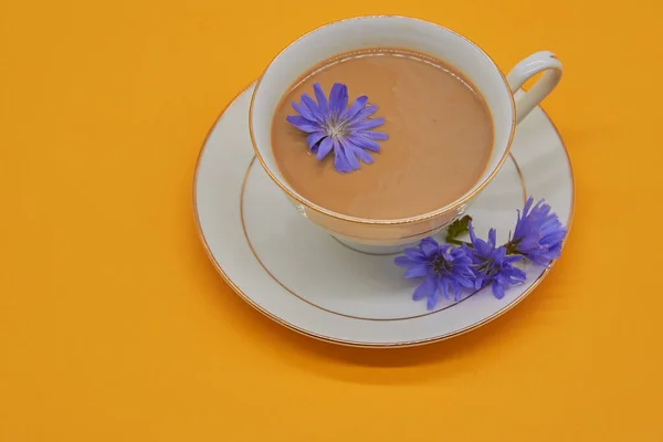 A Cup of chicory drink stands on a saucer on a yellow background, several chicory flowers are lying next to it.