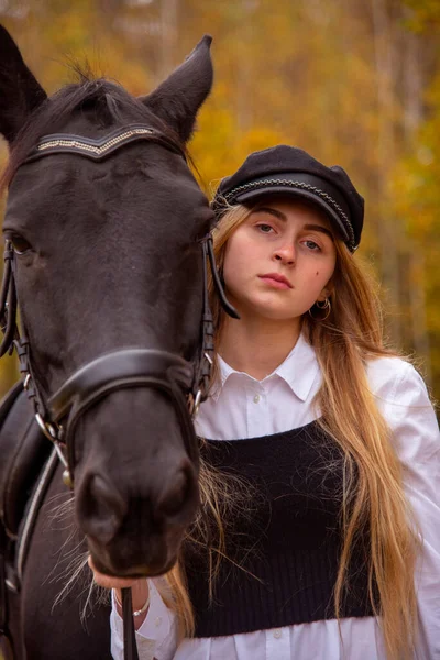 一个金发女郎的画像 头戴一顶帽子 在森林的背景上骑着一匹海湾马 — 图库照片