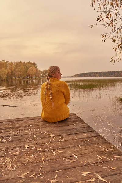 Een tienermeisje zit op de rand van een houten brug over het meer met haar rug naar de camera. — Stockfoto
