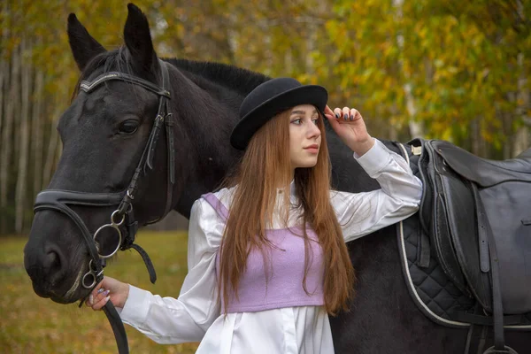 Roodharig meisje met een zwarte hoed en een paard in het herfstbos. — Stockfoto