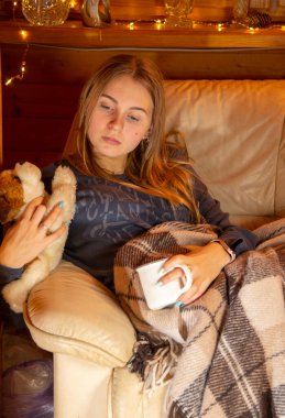 Kız kanepede oturuyor, sıcak bir battaniyeyle örtülü, lambadan gelen ışıkla aydınlatılmış. Ellerinde büyük bir kupa ve yumuşak bir oyuncak var. Arka planda parlak tatil fenerleri.