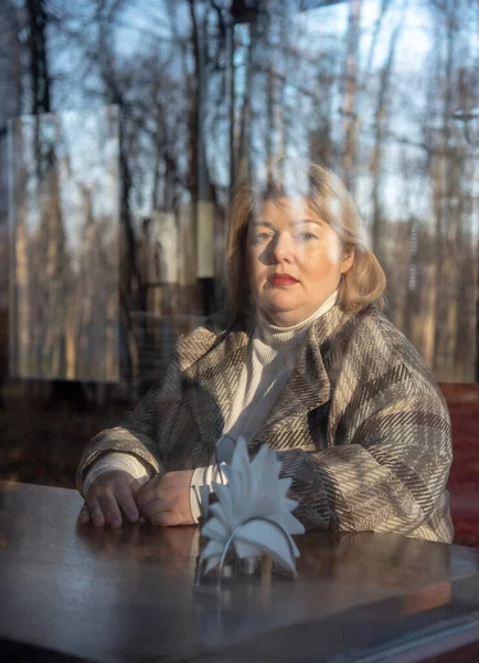 一个中年妇女的画像 一头金发碧眼的中年妇女 穿过咖啡店的窗户 映衬着光秃秃的秋树 — 图库照片