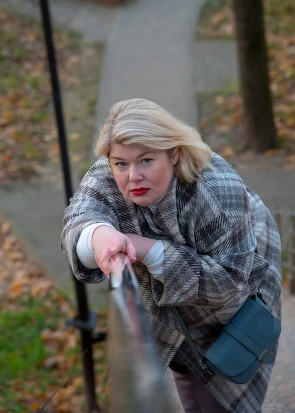 A plump, middle-aged blonde leans on the metal railing on the stairs and looks at the camera. — Stock Photo, Image