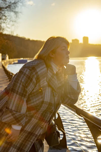 Een mooie mollige vrouw met blond haar en een geruite jas staat op de dijk in de stralen van de ondergaande zon. — Stockfoto