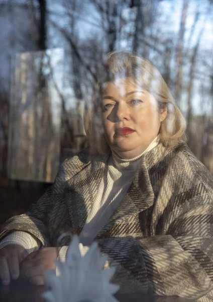 一个胖胖的中年妇女的画像，通过一个咖啡馆的窗户拍摄的，它反映了光秃秃的秋树. — 图库照片