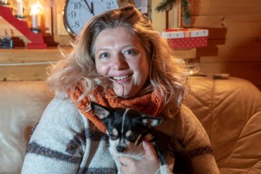 Şişman, orta yaşlı sarışın bir kadın deri koltukta küçük bir köpekle oturuyor, bir chihuahua, noel dekoru olan bir odada..