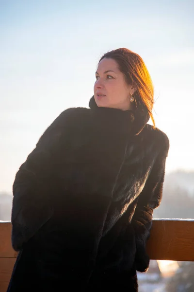 A middle-aged woman with red hair , wearing a black fur coat, against the background of a winter landscape on a sunny day. Portrait in a sunny haze.