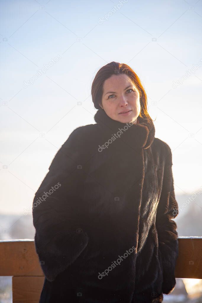 A middle-aged woman with red hair , wearing a black fur coat, against the background of a winter landscape on a sunny day. Portrait in a sunny haze.