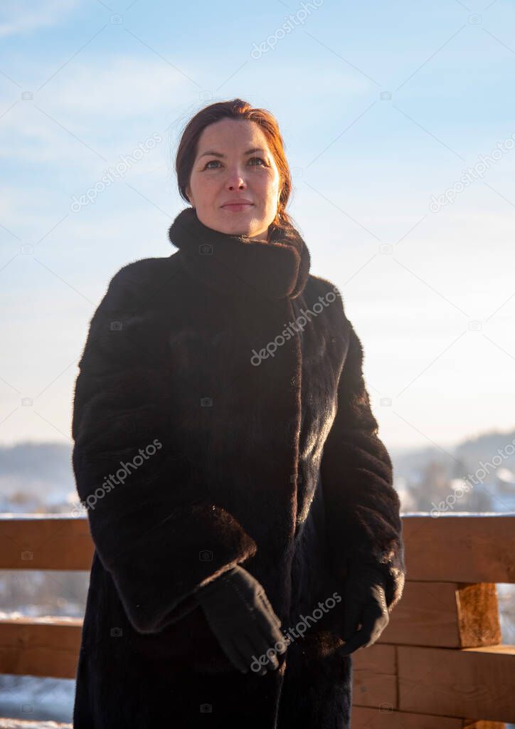 A middle-aged woman , wearing a black fur coat, against the background of a winter landscape on a sunny day.