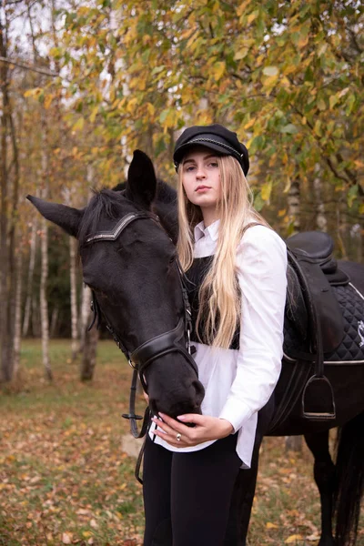 一个有着长长的金发和黑色帽子的少女的画像 她在秋天的森林里抱着一匹栗子马 — 图库照片
