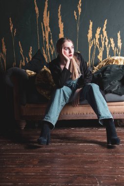 Kotlu ve siyah ceketli bir kız eskimiş bir kanepede, karanlık bir duvarın karşısında oturuyor. Fotoğraf stüdyosunda fotoğraf çekimi..