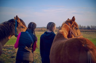 Two teenage girls stand with their backs to the camera and hold the reins of young horses.
