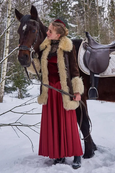 在冬季的森林里 一个金发碧眼 身穿旧式红色连衣裙的女孩站在一匹马旁边 — 图库照片#