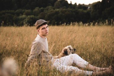 18 yaşında kepli bir çocuk bir köpekle tarlada oturuyor. .