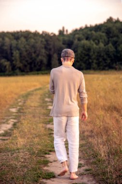 Genç bir adam ormana giden toprak yolda yalınayak yürüyor..
