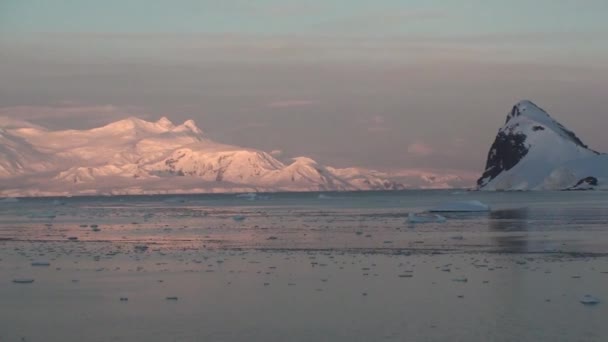 Matahari terbenam di Antartika, dengan air laut yang tenang mencerminkan oranye yang indah dan indah, kuning dan biru dan pegunungan dengan pantai berbatu — Stok Video