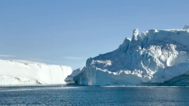Muazzam bir buzdağı sakin sularda görkemli bir şekilde yüzer. Parlak mavi gökyüzü çarpıcı bir zemin sağlıyor, buzdağlarını vurguluyor eşsiz şekiller ve dokular..