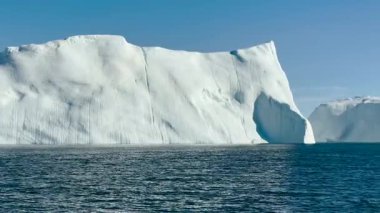 Muazzam bir buzdağı sakin sularda görkemli bir şekilde yüzer. Parlak mavi gökyüzü çarpıcı bir zemin sağlıyor, buzdağlarını vurguluyor eşsiz şekiller ve dokular..