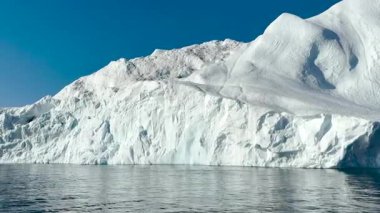 Muazzam bir buzdağı sakin sularda görkemli bir şekilde yüzer. Parlak mavi gökyüzü çarpıcı bir zemin sağlıyor, buzdağlarını vurguluyor eşsiz şekiller ve dokular..