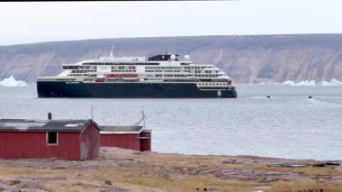Büyük bir yolcu gemisi yüzen buzlarla dolu Kuzey Kutbu sularında süzülüyor. Parlak, güneşli bir gökyüzünün altında arka planda görkemli dağlar yükselir..