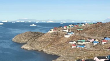 Grönland 'daki deniz kıyısındaki kayalık arazideki canlı evlerin göz kamaştırıcı manzarası. Buzdağları suda yüzer, açık bir günde güzel bir manzara yaratır..