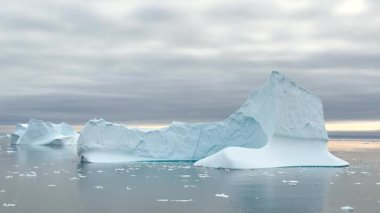 Buzdağları, günbatımının yumuşak parıltısını yansıtan sakin sularda zarifçe sürüklenirler. Kutup sahnesinde sıcak renklerle dolu sakin bir gökyüzüne karşı buz oluşumları sergileniyor..