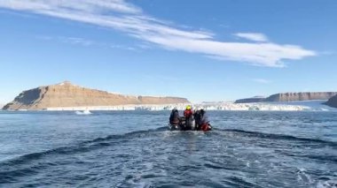 Bir grup maceraperest, çarpıcı bir kutup manzarasındaki yüksek buzdağlarını keşfetmek için şişme bir tekneye biniyor. Su sakin, berrak gökyüzünün altındaki mavi buzu yansıtıyor..