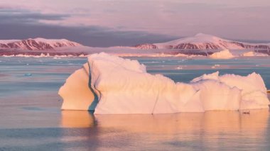 Güneş uzak bir kutup bölgesinin üzerinde batarken buzdağları berrak sularda yavaşça sürüklenir. Ufukta uzak tepeler ve pembe-turuncu bir gökyüzü görünüyor..