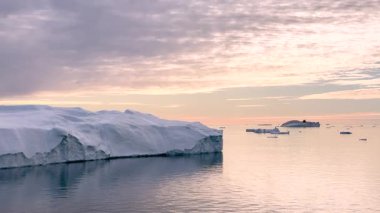 Güneş uzak bir kutup bölgesinin üzerinde batarken buzdağları berrak sularda yavaşça sürüklenir. Ufukta uzak tepeler ve pembe-turuncu bir gökyüzü görünüyor..