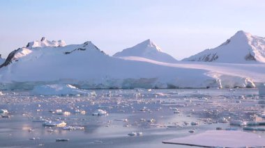 Güneş uzak bir kutup bölgesinin üzerinde batarken buzdağları berrak sularda yavaşça sürüklenir. Ufukta uzak tepeler ve pembe-turuncu bir gökyüzü görünüyor..