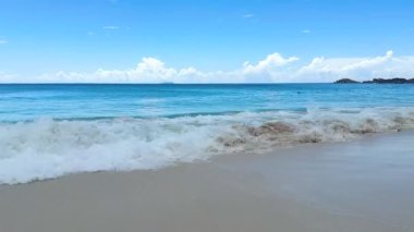 Yumuşak kum ve mavi suyu olan berrak bir sahil uzakta bir tekne gösteriyor. Gökyüzü beyaz bulutlarla aydınlık ve uzak dağlar görülebilir. Ziyaretçiler bu güneşli günün tadını çıkarabilir..