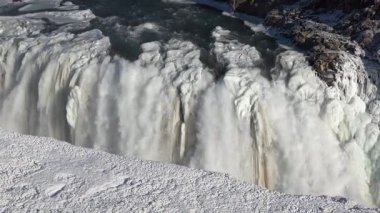 İzlanda 'da Gullfoss şelalesinin kenarında. Parlak güneş ışığı kayalardan aşağı akarken akan suyun üzerinde parlıyor. Manzara, şelalenin etrafındaki manzarayı gösteriyor..