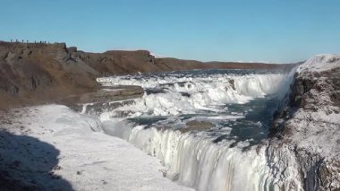 İzlanda 'da Gullfoss şelalesinin kenarında. Parlak güneş ışığı kayalardan aşağı akarken akan suyun üzerinde parlıyor. Manzara, şelalenin etrafındaki manzarayı gösteriyor..
