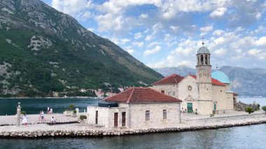 Tarihi binaları ve ağaçları olan bir tekne adaya yaklaşıyor. Gökyüzü bulutlarla kaplıdır ve dağlar bir zemin oluşturur. İnsanlar sahilde günün tadını çıkarırken görülebilir..