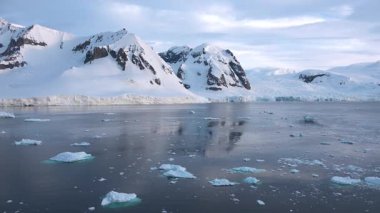 Buzdağları, arka planda duran yüksek dağlar gibi buzlu sularda sürükleniyor. Gökyüzü bulutlu ve manzara soğuk bir günde Antarktika 'nın güzelliğini gösteriyor..