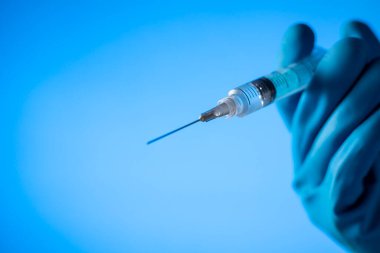 Elleri mavi eldivenli, elinde aşı olan bir şırınga var. Covid-19 koronavirüs. mavi arkaplanda.