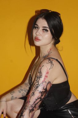 Stüdyoda sarı arka planda duran dövmeli genç bir kadın.