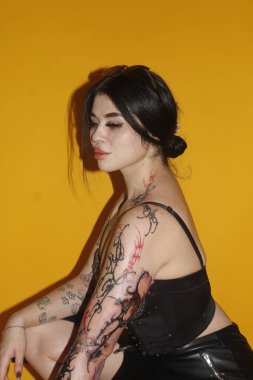 Stüdyoda sarı arka planda duran dövmeli genç bir kadın.