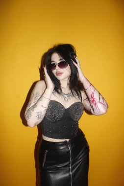 Siyah deri pantolonlu, güneş gözlüklü, sarı arka planda poz veren güzel bir kadın.