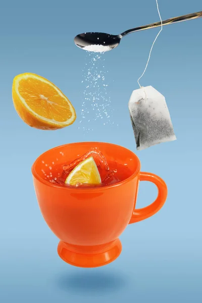 Levitating orange teacup with spoon, tea bag and lemon over the blue surface
