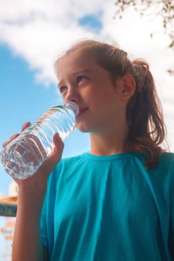 Mavi gömlekli bir kız şişeden su içiyor.
